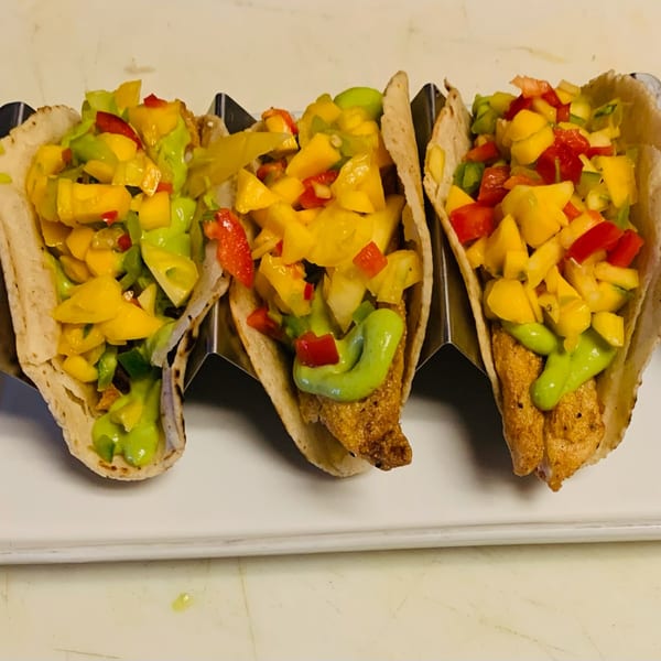 Three fish tacos with mango salsa and green sauce in a metal holder on a white plate.