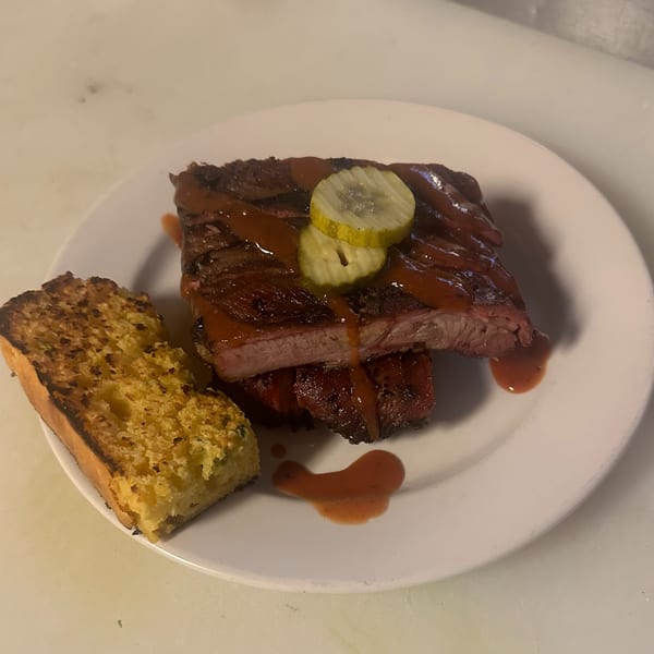 Barbecue ribs with sauce and pickle slices, served with a side of cornbread on a white plate.