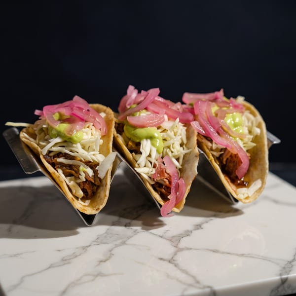 Three tacos in a metal holder, topped with shredded cabbage, green sauce, and pickled red onions.